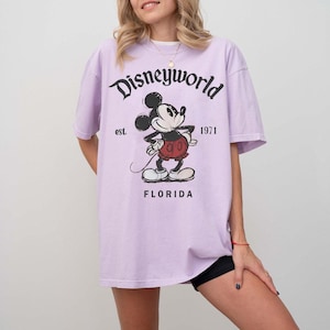May include: A light purple t-shirt featuring a graphic of Mickey Mouse with the text "Disneyworld" above and "Florida" below. The shirt also has the text "est. 1971".