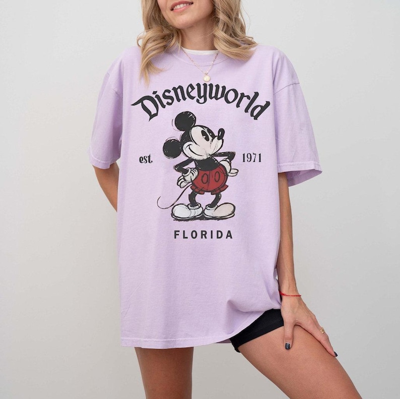 May include: A light purple t-shirt featuring a graphic of Mickey Mouse with the text "Disneyworld" above and "Florida" below. The shirt also has the text "est. 1971".