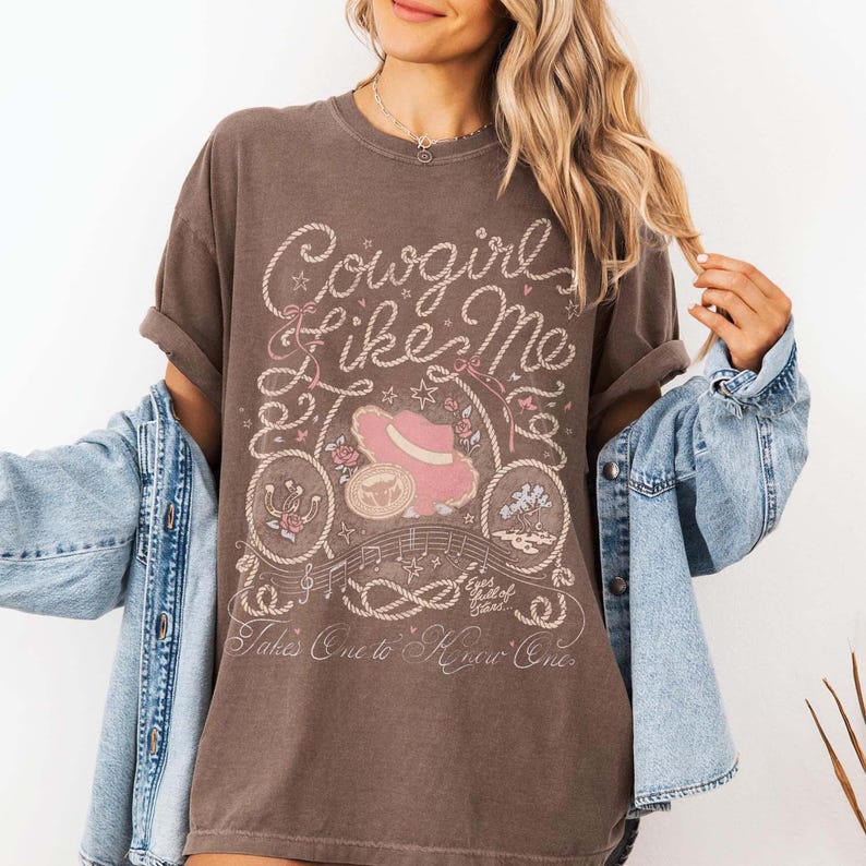 May include: A dark brown short-sleeve t-shirt featuring a graphic print with the text Cowgirl Like Me and Takes One to Know One. The design includes a pink hat, rope borders, stars, musical notes, and a horseshoe in light pink and white ink.