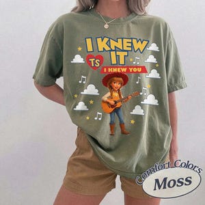 May include: An olive green short-sleeve t-shirt featuring a graphic of a cowgirl playing an acoustic guitar. The text reads I KNEW IT and I KNEW YOU, accompanied by a red heart, white clouds, yellow stars, and musical notes in a cartoon style.