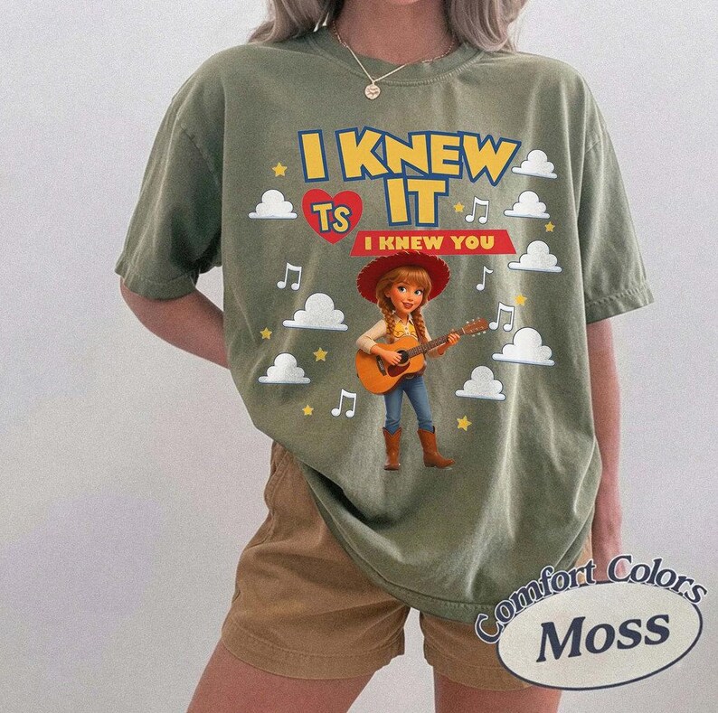 May include: An olive green short-sleeve t-shirt featuring a graphic of a cowgirl playing an acoustic guitar. The text reads I KNEW IT and I KNEW YOU, accompanied by a red heart, white clouds, yellow stars, and musical notes in a cartoon style.