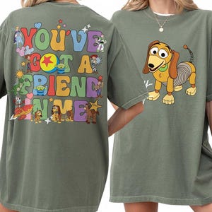 May include: An olive green t-shirt featuring a graphic on the back with the text "YOU'VE GOT A FRIEND IN ME" surrounded by various cartoon characters and colorful shapes. The front of the shirt displays a graphic of a brown and tan toy dog.