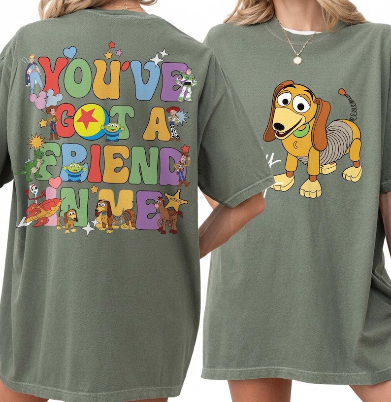 May include: An olive green t-shirt featuring a graphic on the back with the text "YOU'VE GOT A FRIEND IN ME" surrounded by various cartoon characters and colorful shapes. The front of the shirt displays a graphic of a brown and tan toy dog.