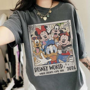 May include: Dark gray t-shirt featuring a colorful graphic of Disney characters including Mickey Mouse, Minnie Mouse, Donald Duck, and Goofy. The graphic reads "Disney World 2026" with the tagline "Where Dreams Come True."
