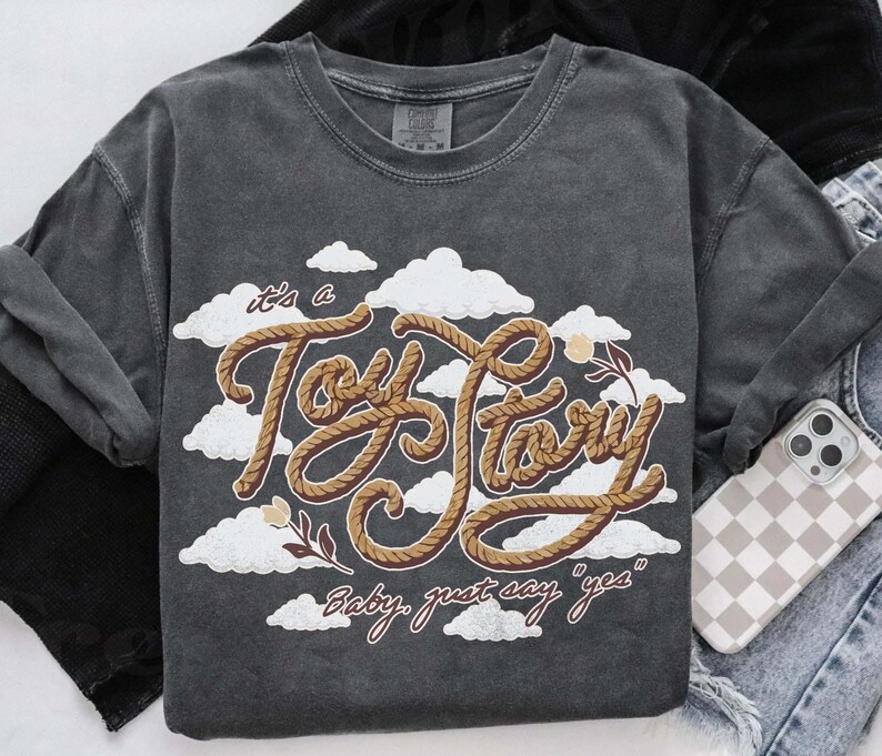 May include: A charcoal gray short-sleeve t-shirt featuring a graphic of white clouds and the text "it's a Toy Story" written in a brown rope-style font, with the additional text "Baby, just say "yes"" printed below in a brown cursive script.