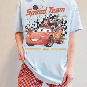 May include: Light blue t-shirt featuring a graphic of Lightning McQueen from the Cars movie with the text "Speed Team 95 Cars 1928". The shirt also features Mickey Mouse, Minnie Mouse, Goofy, and Donald Duck.