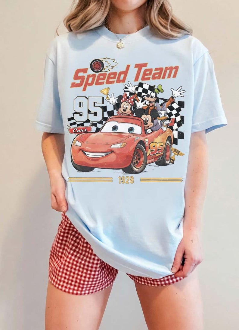 May include: Light blue t-shirt featuring a graphic of Lightning McQueen from the Cars movie with the text "Speed Team 95 Cars 1928". The shirt also features Mickey Mouse, Minnie Mouse, Goofy, and Donald Duck.