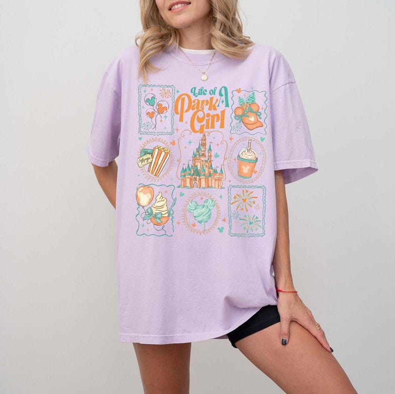 May include: A light purple t-shirt with the text "Life of a Park Girl" and various illustrations. The illustrations include a castle, popcorn, ice cream, and other park-themed images. The shirt is a relaxed fit.