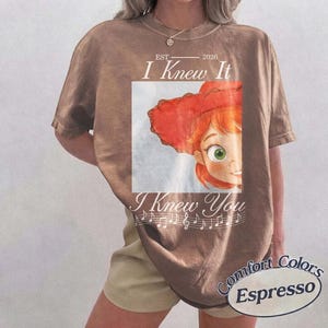 Peut inclure: Un t-shirt marron &agrave; manches courtes avec un graphique repr&eacute;sentant un personnage aux cheveux roux portant un chapeau rouge. Le texte indique "EST 2026 I Knew It I Knew You" avec des notes de musique. Le t-shirt a un col rond et une coupe d&eacute;contract&eacute;e.