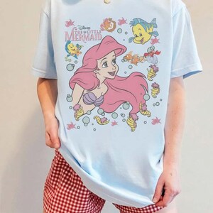 May include: Light blue t-shirt featuring a graphic of Ariel from Disney's The Little Mermaid. The design includes Ariel with pink hair, sea creatures, and the text "Disney The Little Mermaid". The shirt is a casual wear item.