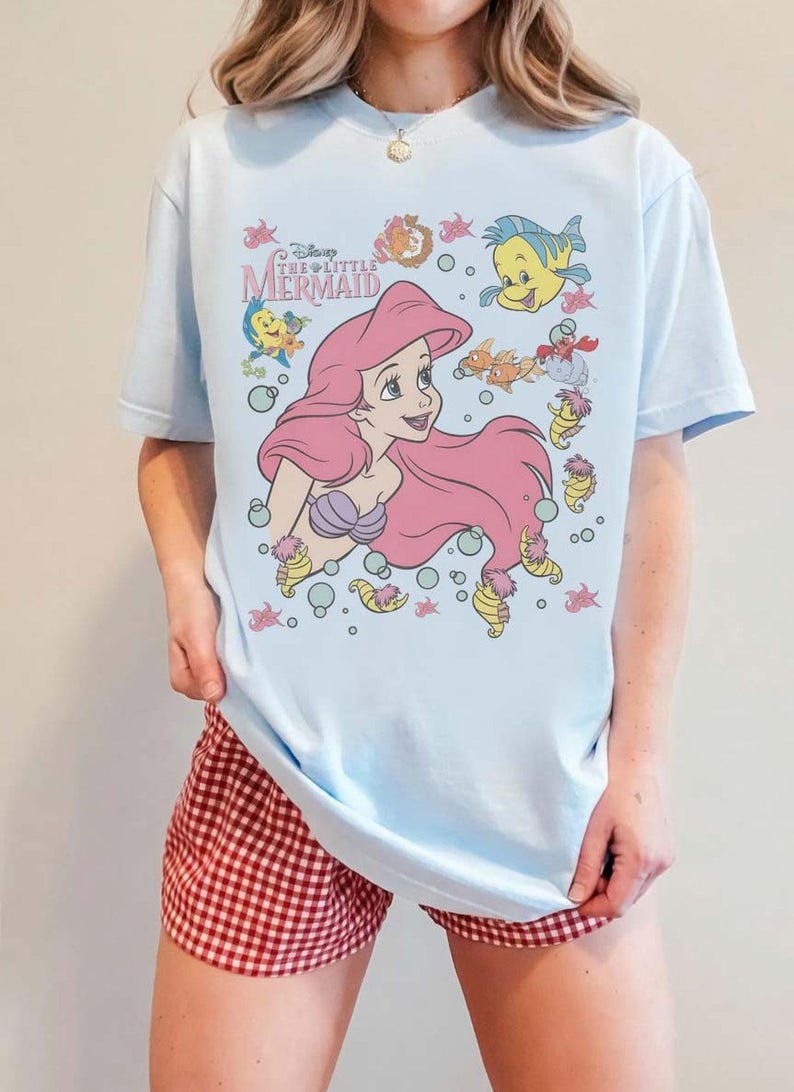 May include: Light blue t-shirt featuring a graphic of Ariel from Disney's The Little Mermaid. The design includes Ariel with pink hair, sea creatures, and the text "Disney The Little Mermaid". The shirt is a casual wear item.