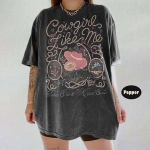 May include: A charcoal gray short-sleeve t-shirt featuring a graphic print with the text Cowgirl Like Me and Takes One to Know One. The design includes a pink cowboy hat, rope motifs, musical notes, stars, and floral accents in a vintage style.