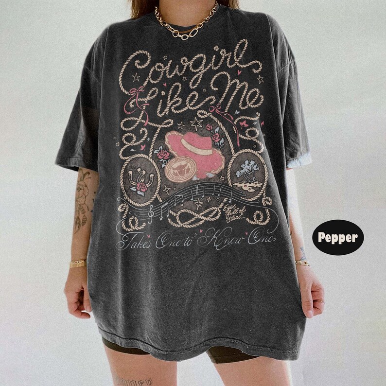 May include: A charcoal gray short-sleeve t-shirt featuring a graphic print with the text Cowgirl Like Me and Takes One to Know One. The design includes a pink cowboy hat, rope motifs, musical notes, stars, and floral accents in a vintage style.