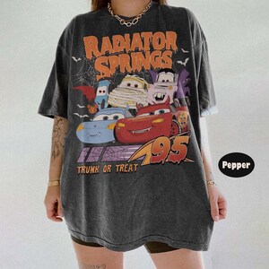 May include: Dark gray t-shirt featuring a graphic of characters from the movie Cars, with the text "Radiator Springs" in orange and "Trunk or Treat" below. The design includes bats and a spiderweb, with the number 95 in orange.