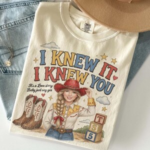 May include: A cream-colored t-shirt featuring a graphic of a person with a blonde braid wearing a red cowboy hat and shirt. The text reads I KNEW IT. I KNEW YOU. It's a Love story Baby just say yes. Includes illustrations of cowboy boots, clouds, stars, and three stacked alphabet blocks.
