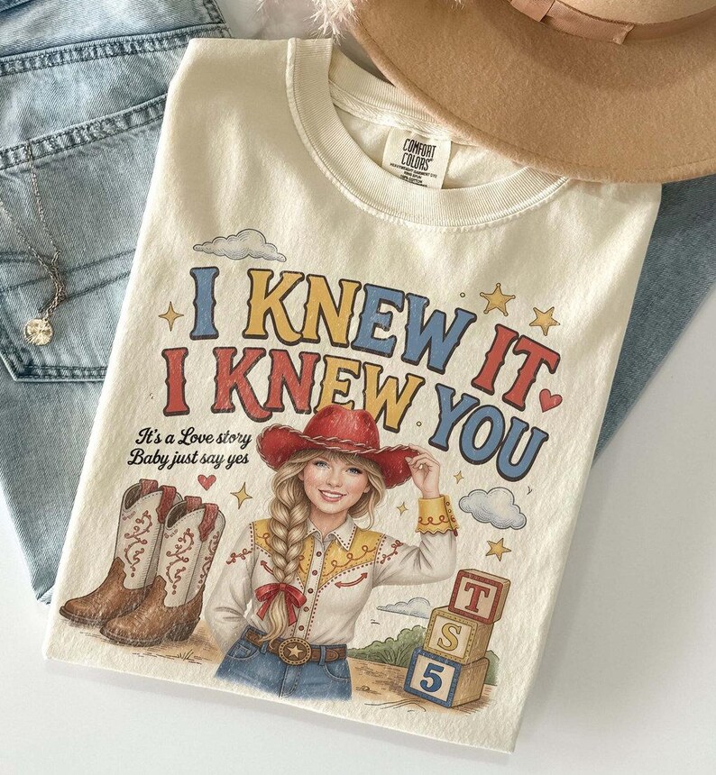 May include: A cream-colored t-shirt featuring a graphic of a person with a blonde braid wearing a red cowboy hat and shirt. The text reads I KNEW IT. I KNEW YOU. It's a Love story Baby just say yes. Includes illustrations of cowboy boots, clouds, stars, and three stacked alphabet blocks.