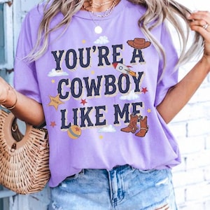 以下が含まれることがあります： 「YOU'RE A COWBOY LIKE ME」というダークブルーの文字がプリントされた、ライトパープルの半袖Tシャツ。グラフィックには、茶色のカウボーイハット、ロケット、TAYLORと書かれた黄色い保安官の星、ボール、小さな星が描かれています。