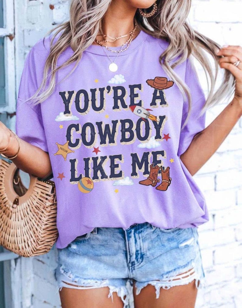 以下が含まれることがあります： 「YOU'RE A COWBOY LIKE ME」というダークブルーの文字がプリントされた、ライトパープルの半袖Tシャツ。グラフィックには、茶色のカウボーイハット、ロケット、TAYLORと書かれた黄色い保安官の星、ボール、小さな星が描かれています。