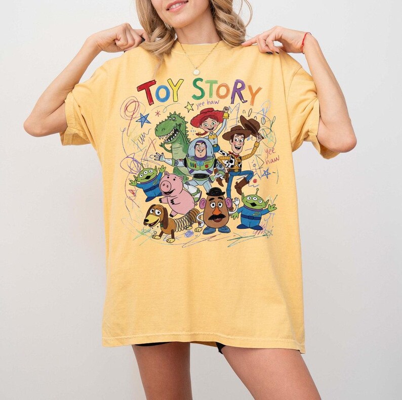 May include: Yellow t-shirt featuring the words "TOY STORY" in colorful letters with cartoon characters. The shirt has a crew neck and short sleeves. The design includes Woody, Buzz Lightyear, and other characters.