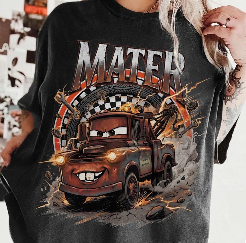 Pu&ograve; includere: T-shirt nera con un disegno di un carro attrezzi dei cartoni animati, con la scritta "MATER" sopra. Il design include strumenti, fulmini e una bandiera a scacchi, creando un tema di corse.