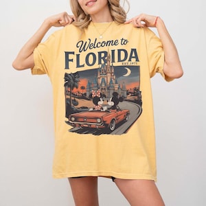 May include: Mustard yellow t-shirt with a graphic that says "Welcome to Florida" and features a vintage car with Mickey and Minnie Mouse. The graphic also includes a castle, palm trees, and a crescent moon.
