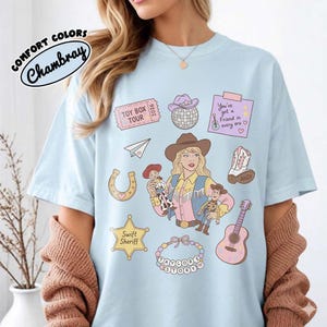May include: A light blue t-shirt featuring a graphic print of a blonde person in a cowboy hat holding two dolls, surrounded by illustrations of a disco ball, a guitar, a horseshoe, a sheriff badge, a paper airplane, and a beaded necklace.