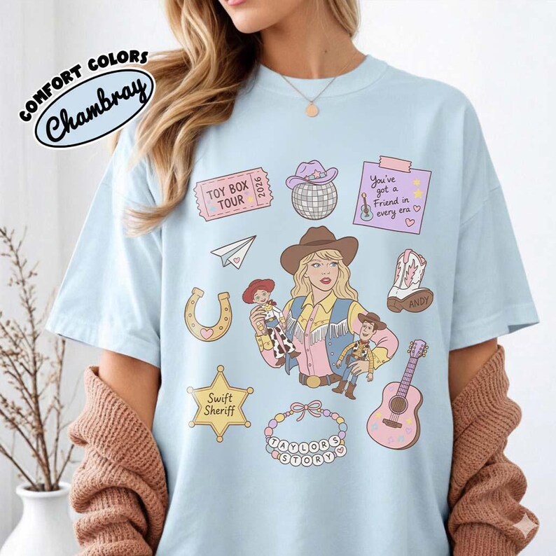 May include: A light blue t-shirt featuring a graphic print of a blonde person in a cowboy hat holding two dolls, surrounded by illustrations of a disco ball, a guitar, a horseshoe, a sheriff badge, a paper airplane, and a beaded necklace.