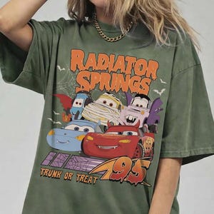 May include: Green t-shirt featuring a Halloween-themed graphic of the Cars characters. The design includes the text "Radiator Springs" and "Trunk or Treat 95". The graphic features Lightning McQueen and other characters in costumes.