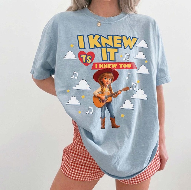 May include: A light blue t-shirt featuring a graphic of a cowgirl playing an acoustic guitar. The text reads I KNEW IT and I KNEW YOU, accompanied by a red heart, yellow stars, white clouds, and musical notes printed on the front of the garment.