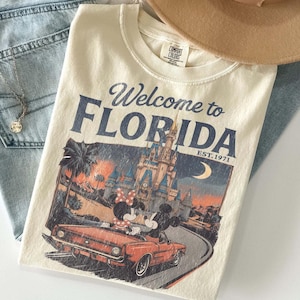 May include: Cream-colored t-shirt with the text "Welcome to Florida" and a graphic of Mickey and Minnie Mouse in a convertible in front of a castle. The shirt also has the text "EST. 1971".