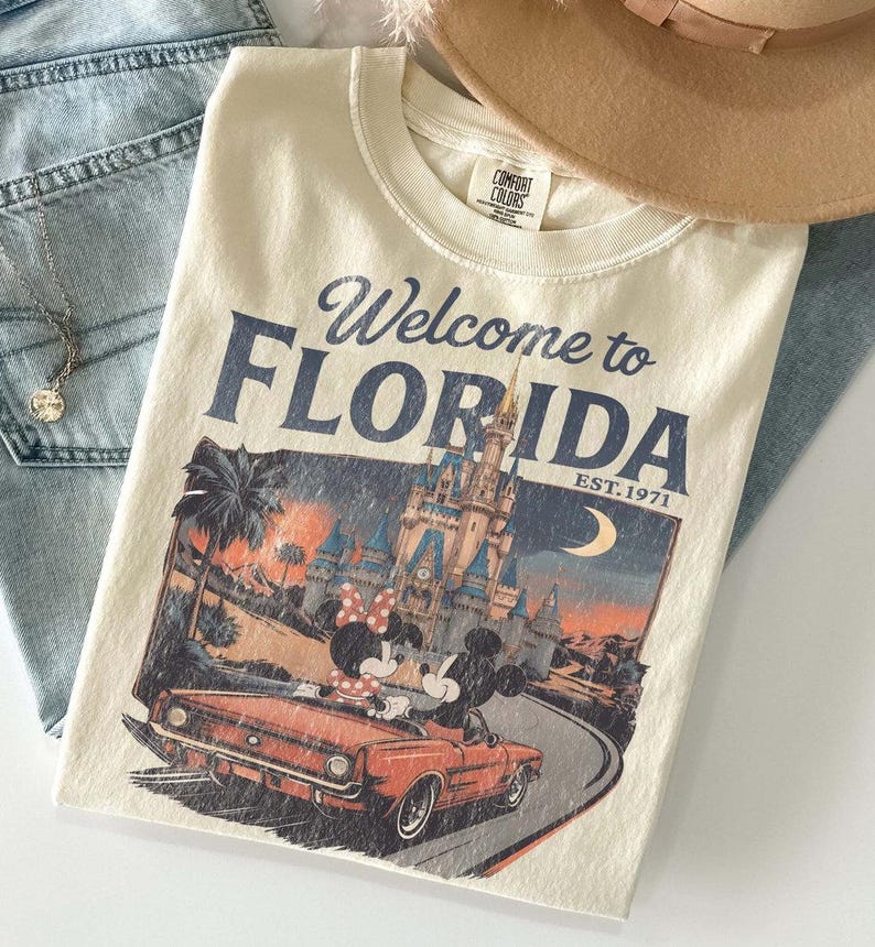 May include: Cream-colored t-shirt with the text "Welcome to Florida" and a graphic of Mickey and Minnie Mouse in a convertible in front of a castle. The shirt also has the text "EST. 1971".