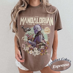 May include: Espresso-colored t-shirt featuring a graphic of The Mandalorian and Grogu surrounded by flowers. The text "THE MANDALORIAN - THIS IS THE WAY" is printed above the image. The shirt is a Comfort Colors brand.