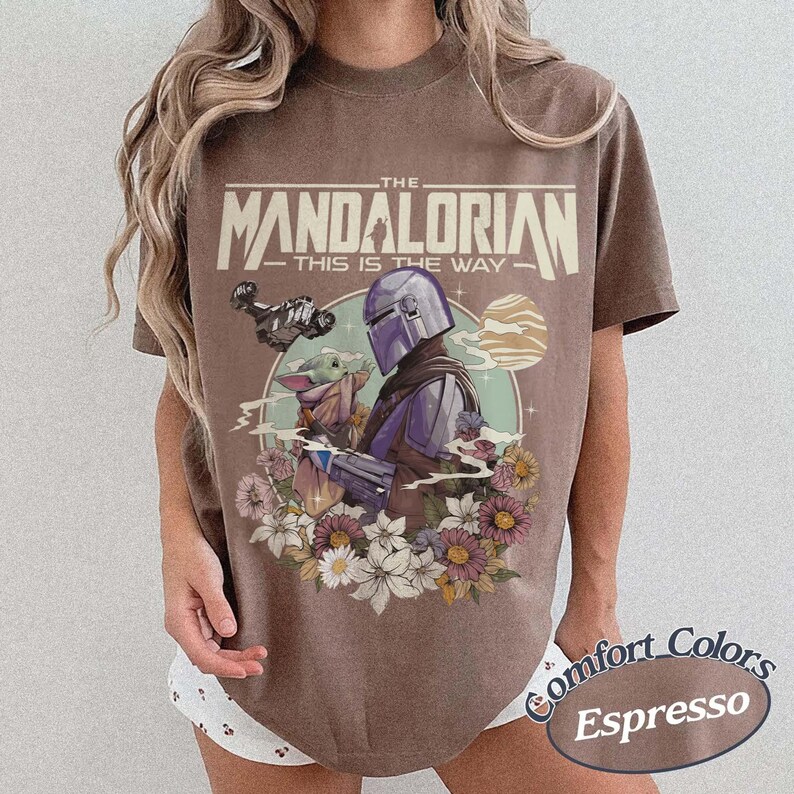 May include: Espresso-colored t-shirt featuring a graphic of The Mandalorian and Grogu surrounded by flowers. The text "THE MANDALORIAN - THIS IS THE WAY" is printed above the image. The shirt is a Comfort Colors brand.