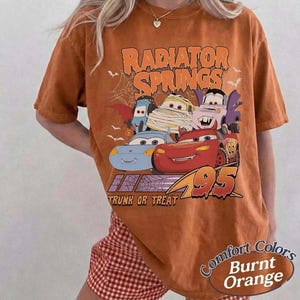 May include: Burnt orange t-shirt featuring a graphic of cartoon cars from Radiator Springs, with the text "Radiator Springs" and "Trunk or Treat". The shirt also has the text "Comfort Colors" and "Burnt Orange".