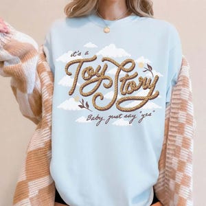 May include: A light blue crewneck sweatshirt featuring a graphic of white clouds and the text "it's a Toy Story" written in a brown rope-style font, with the additional script text "Baby, just say "yes"" printed below in a dark brown color.
