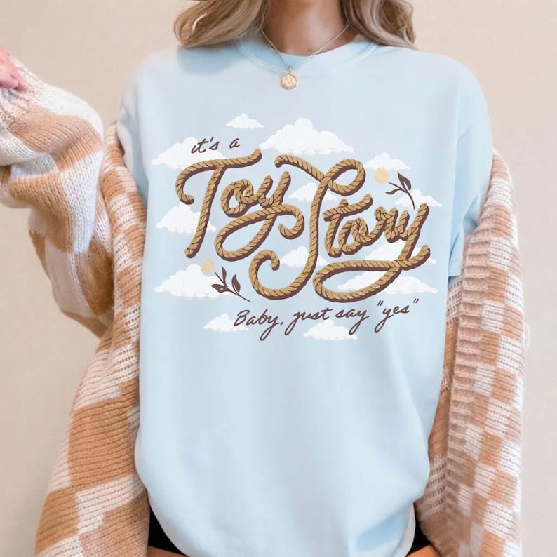 May include: A light blue crewneck sweatshirt featuring a graphic of white clouds and the text "it's a Toy Story" written in a brown rope-style font, with the additional script text "Baby, just say "yes"" printed below in a dark brown color.