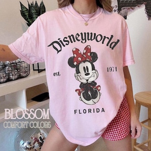 May include: A light pink t-shirt with the text "Disneyworld" above a Minnie Mouse graphic. The graphic includes the text "est. 1971" and "FLORIDA". The shirt also has the text "BLOSSOM COMFORT COLORS".