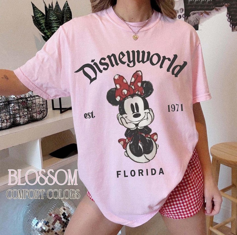 May include: A light pink t-shirt with the text "Disneyworld" above a Minnie Mouse graphic. The graphic includes the text "est. 1971" and "FLORIDA". The shirt also has the text "BLOSSOM COMFORT COLORS".