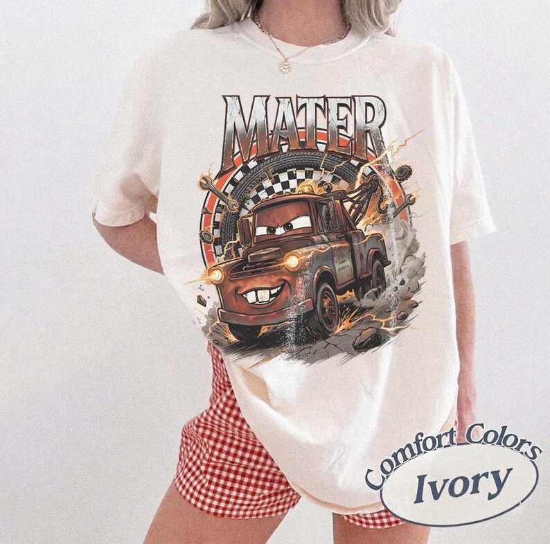 Pu&ograve; includere: T-shirt color avorio con un'immagine a cartoni animati di Mater del film Cars. La grafica include la parola "MATER", una bandiera a scacchi e fulmini. La maglietta &egrave; etichettata "Comfort Colors Ivory".