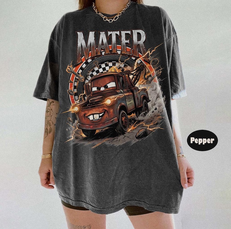 Pu&ograve; includere: T-shirt grigio scuro con una grafica invecchiata di Cricchetto del film Cars. Il design include la parola "MATER" in grassetto sopra il personaggio, con uno sfondo a tema racing. La maglietta ha maniche corte e una vestibilit&agrave; ampia.