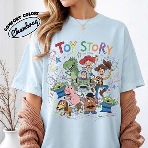 May include: Light blue t-shirt featuring the words "TOY STORY" in colorful letters, with cartoon characters from the movie. The shirt has the words "Comfort Colors Chambray" in a circular design.