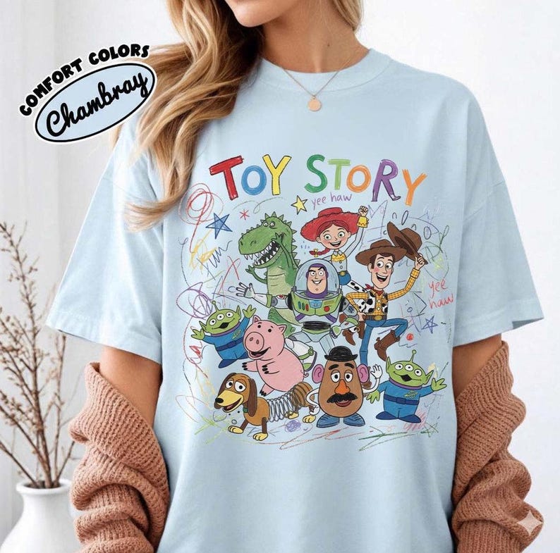 May include: Light blue t-shirt featuring the words "TOY STORY" in colorful letters, with cartoon characters from the movie. The shirt has the words "Comfort Colors Chambray" in a circular design.