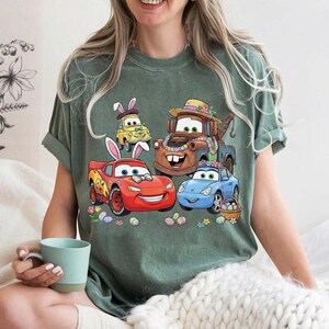 Pode incluir: T-shirt verde com um gr&aacute;fico colorido de carros animados com orelhas de coelho da P&aacute;scoa e decora&ccedil;&otilde;es florais. O design inclui um carro vermelho, um carro azul, um carro amarelo e um caminh&atilde;o marrom, junto com ovos de P&aacute;scoa e flores.