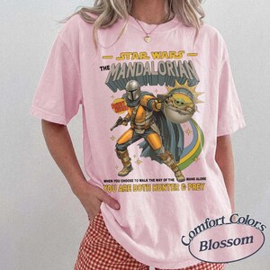 May include: A light pink short-sleeve t-shirt featuring a graphic of The Mandalorian and Grogu. The text reads Star Wars The Mandalorian and When You Choose To Walk The Way Of The Mandalore You Are Both Hunter And Prey in yellow and black.