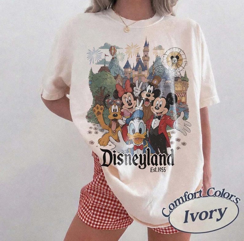 Pu&ograve; includere: Una t-shirt color avorio con una stampa grafica colorata di Topolino, Minnie, Paperino, Pippo e Pluto davanti a un castello. Il testo recita Disneyland Est. 1955. La maglietta ha maniche corte e un girocollo.