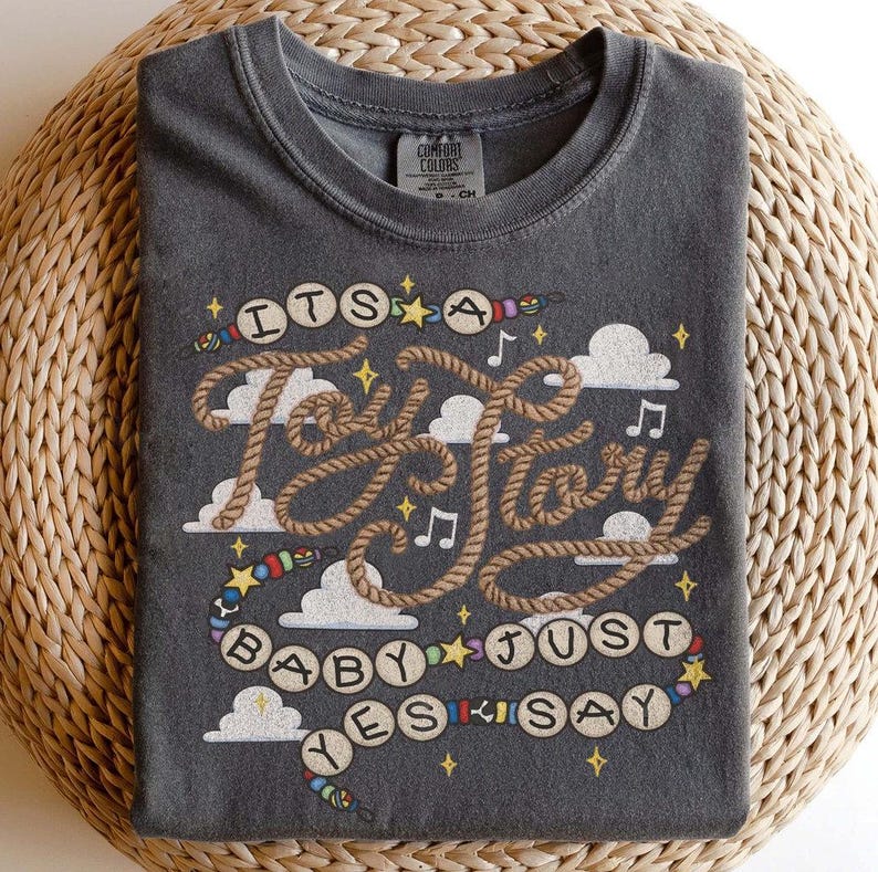 May include: A charcoal gray crew neck t-shirt featuring a graphic print with the text "ITS A Toy Story BABY JUST YES SAY". The design includes rope-style lettering, white clouds, musical notes, small yellow stars, and colorful beads on strings.