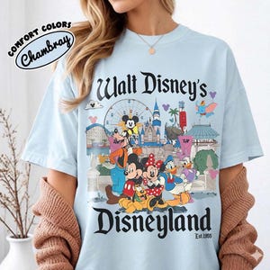 Puede incluir: Camiseta azul claro con un gr&aacute;fico colorido de Disneyland con Mickey Mouse, Minnie Mouse y otros personajes de Disney. La camiseta tiene el texto "Walt Disney's Disneyland" y "Est. 1955". Hecha de material Comfort Colors Chambray.