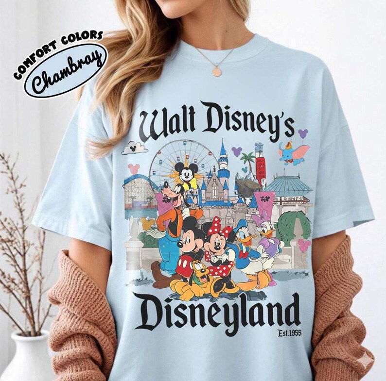 Puede incluir: Camiseta azul claro con un gr&aacute;fico colorido de Disneyland con Mickey Mouse, Minnie Mouse y otros personajes de Disney. La camiseta tiene el texto "Walt Disney's Disneyland" y "Est. 1955". Hecha de material Comfort Colors Chambray.