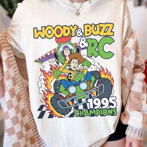 May include: Cream-colored t-shirt featuring a graphic of Woody, Buzz Lightyear, and RC from Toy Story. The design includes the text "WOODY & BUZZ & RC 1995 CHAMPIONS" in yellow and green. The characters are depicted on a green race car with flames.