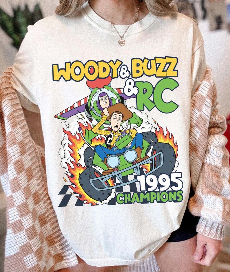 May include: Cream-colored t-shirt featuring a graphic of Woody, Buzz Lightyear, and RC from Toy Story. The design includes the text "WOODY & BUZZ & RC 1995 CHAMPIONS" in yellow and green. The characters are depicted on a green race car with flames.